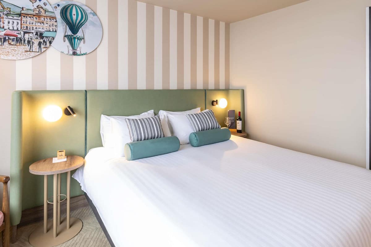 Una habitación de hotel con cama, pared a rayas, mesita de noche redonda y una lámpara de pared.