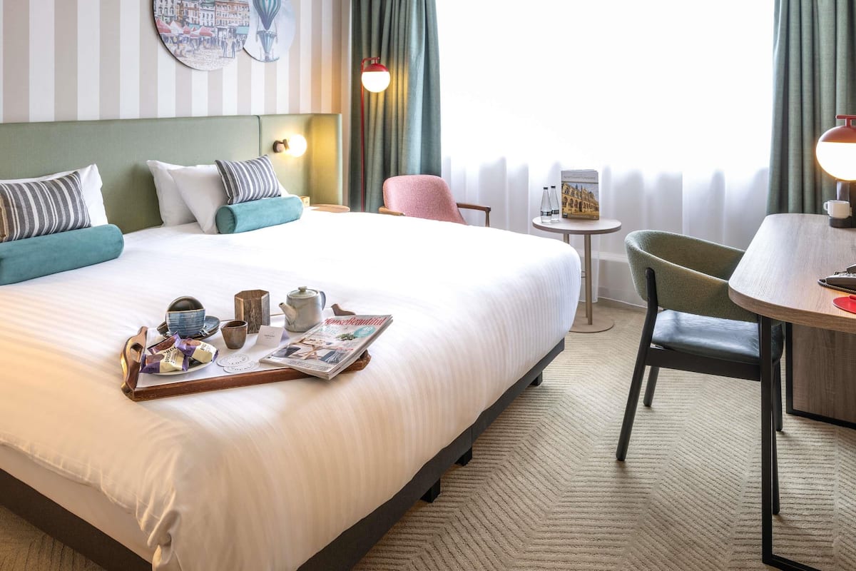 Una habitación de hotel con una cama grande, una mesita, una silla, una lámpara y una ventana con cortinas.
