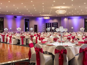 A banquet hall with round tables, white chairs with red sashes, and crystal chandeliers. - Mercure Northampton (Northampton)