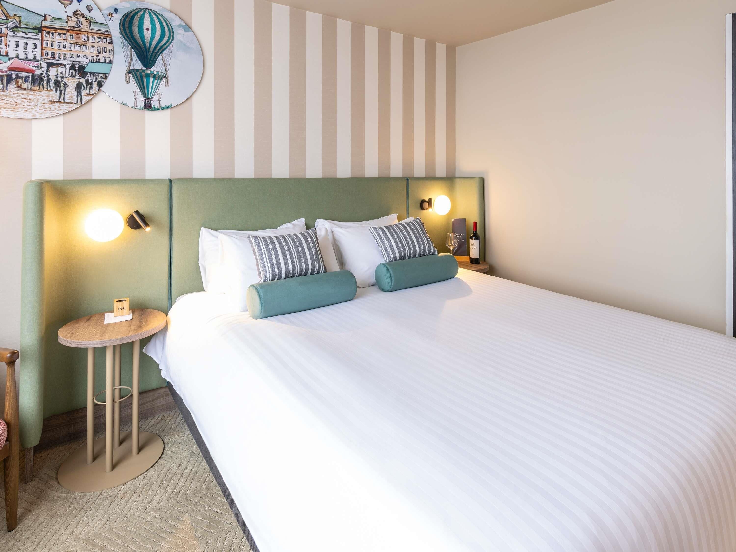 A hotel room with a bed, striped wall, round bedside table, and a wall-mounted light.