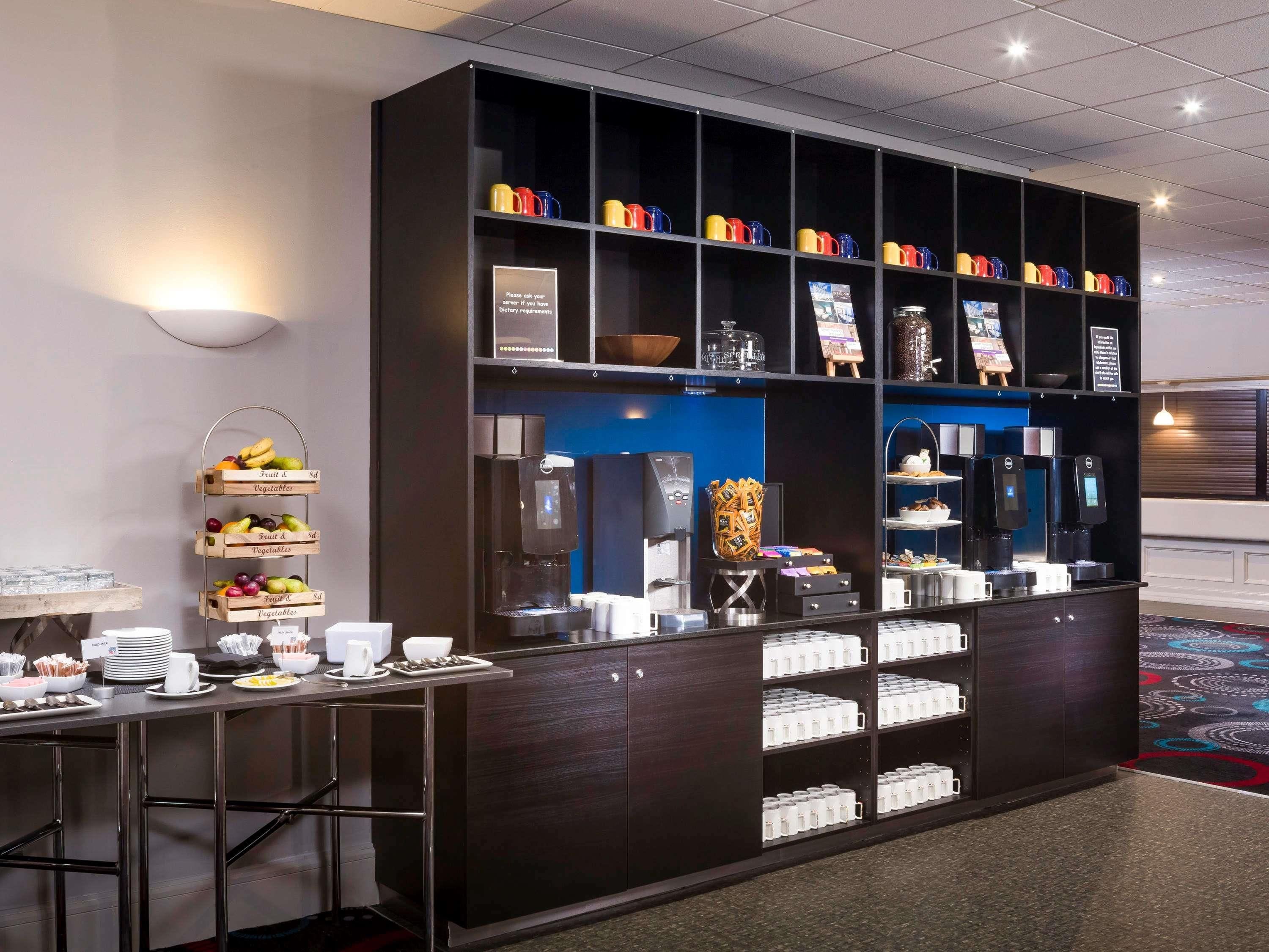 A hotel reception area with a coffee station, fruit and vegetable display, and a digital kiosk.