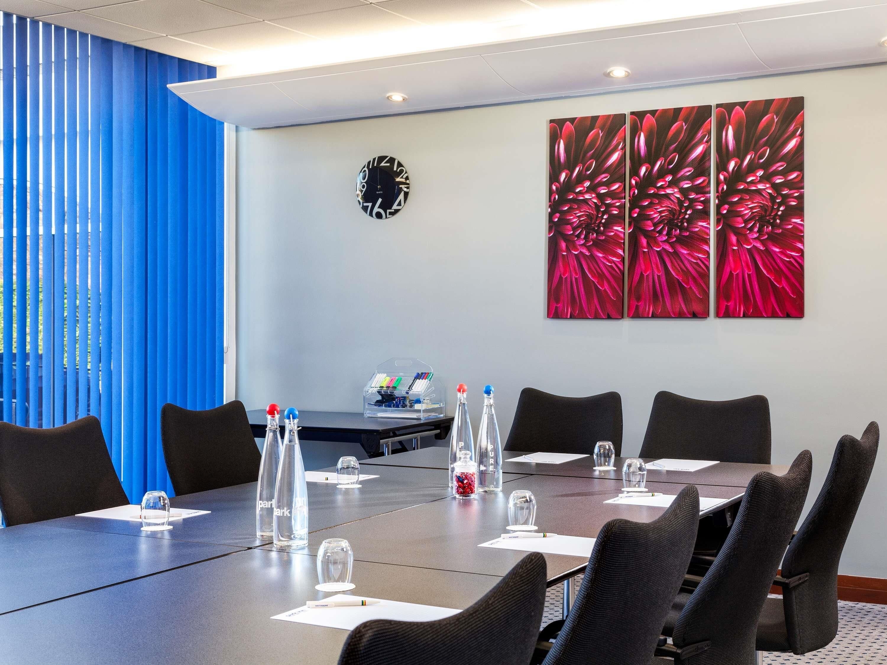 Un salón de conferencias con una larga mesa, sillas, botellas de agua, vasos, un reloj y un cuadro con flores en la pared.