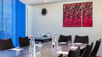 Un salón de conferencias con una larga mesa, sillas, botellas de agua, vasos, un reloj y un cuadro con flores en la pared.