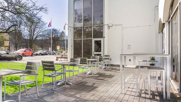 A covered outdoor seating area with tables and chairs, a flag, and a building in the background.