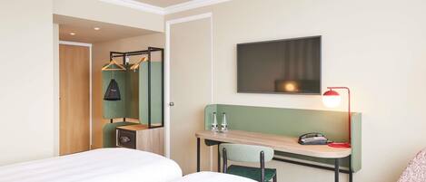 A hotel room with a bed, a desk with a chair, a hanging coat rack, a mirror, a television, and a red lamp.