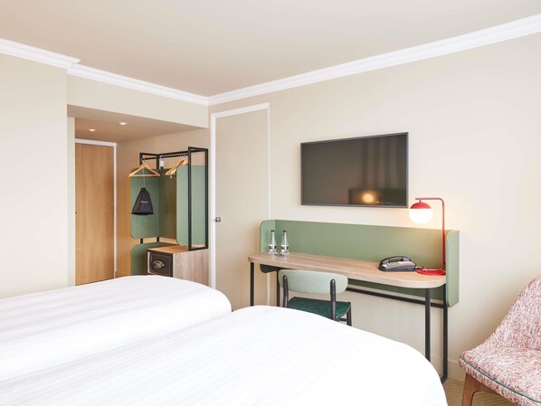 A hotel room with a bed, a desk with a chair, a hanging coat rack, a mirror, a television, and a red lamp.