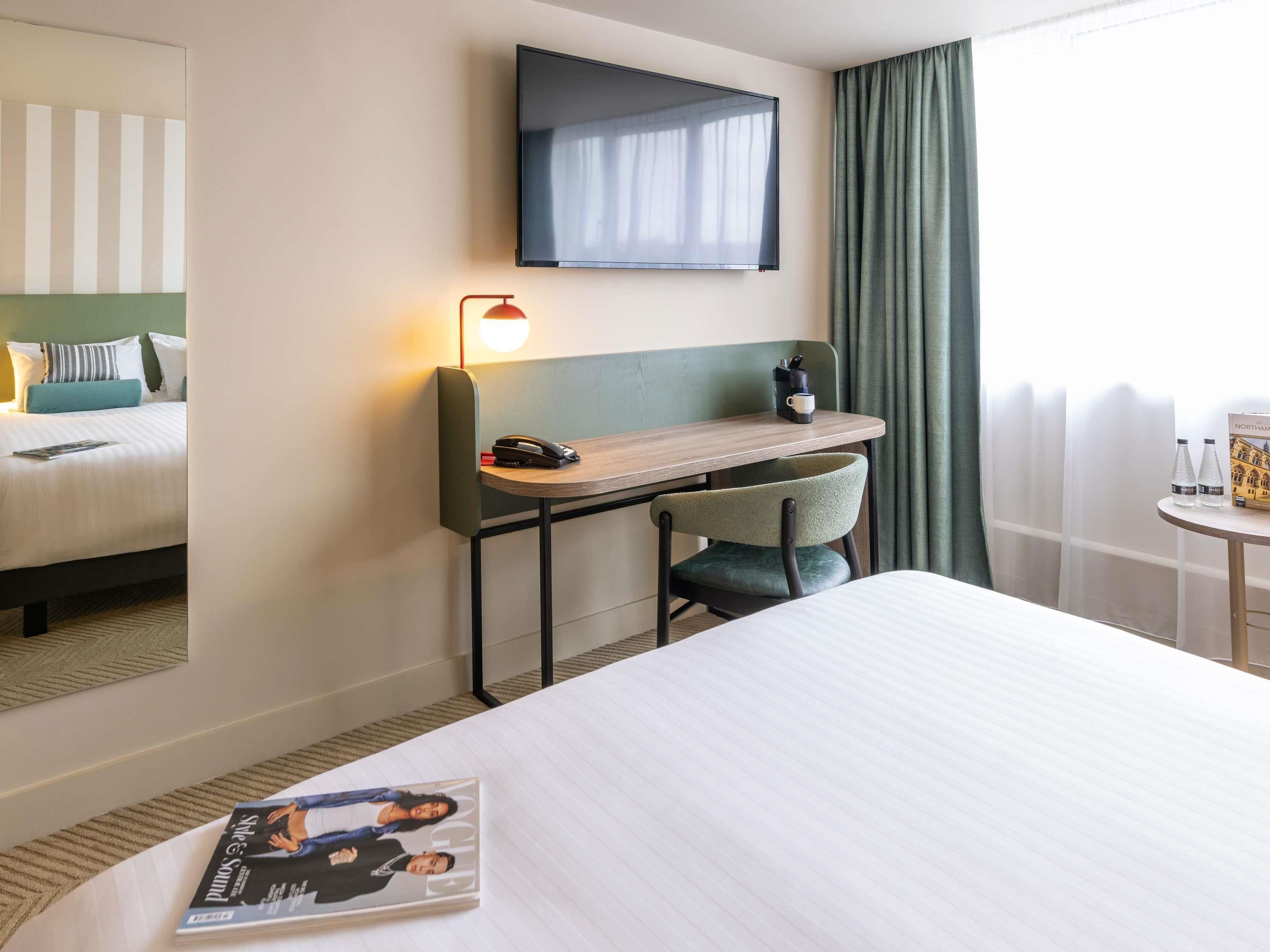 A hotel room with a bed, a desk with a chair, a TV, and a window with curtains.