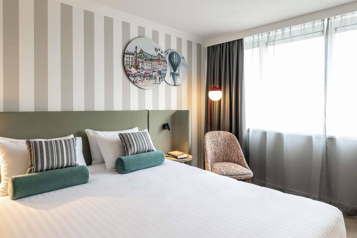 Habitación de hotel con una cama grande, decoración en la pared a rayas, una silla y una ventana con cortinas.