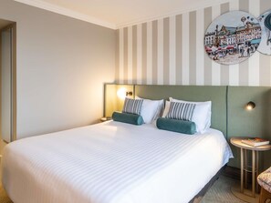 Une chambre d’hôtel avec un grand lit, un papier peint à rayures, une horloge ronde accrochée au mur et une petite table sur laquelle se trouve un livre.