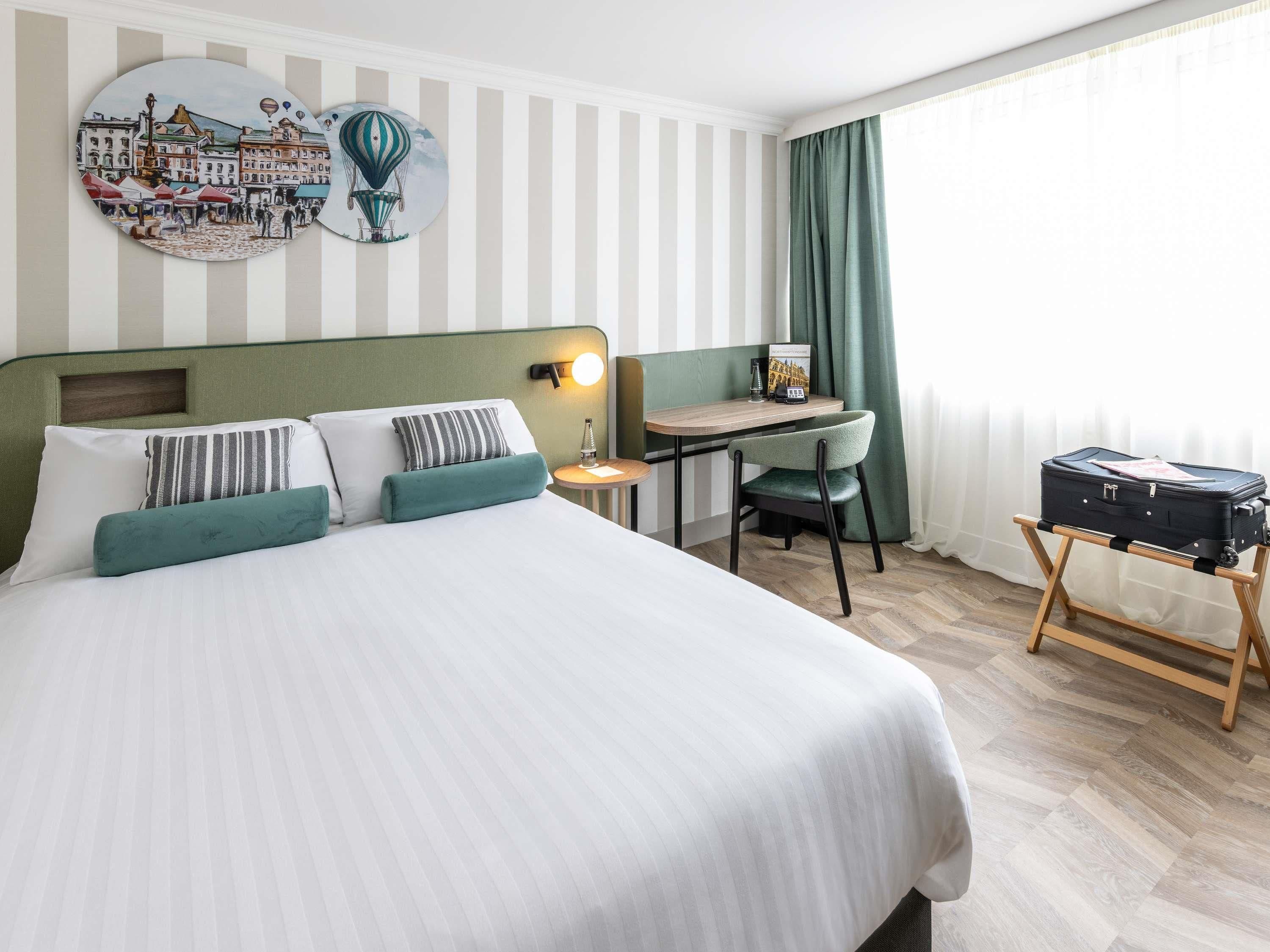 A hotel room with a bed, a desk, a chair, and a suitcase on a stand.