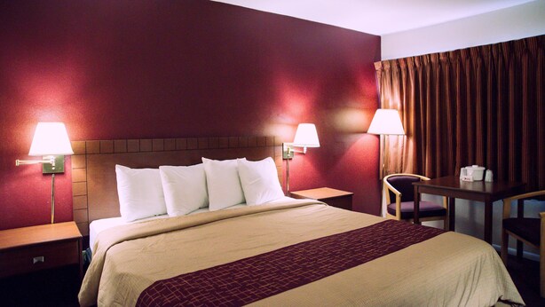 A hotel room with a large bed, two bedside lamps, a wooden nightstand, a small table with a chair, and a curtain on the window. - Red Roof Inn Blythe (Blythe)