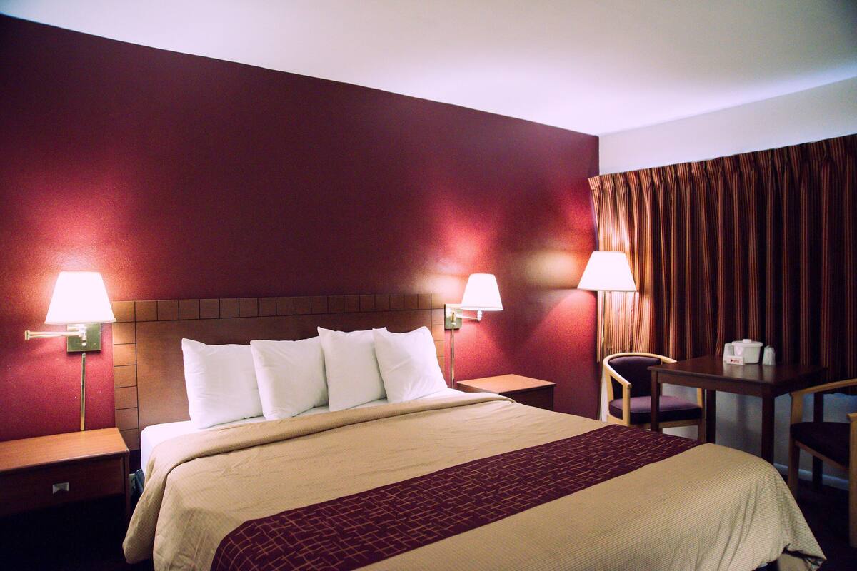 Ein Hotelzimmer mit einem großen Bett, zwei Nachttischlampen, einem hölzernen Nachttisch, einem kleinen Tisch mit Stuhl und einem Vorhang am Fenster.