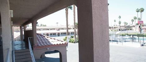 A covered outdoor area with a pool, palm trees, and a building with a red-tiled roof in the background.