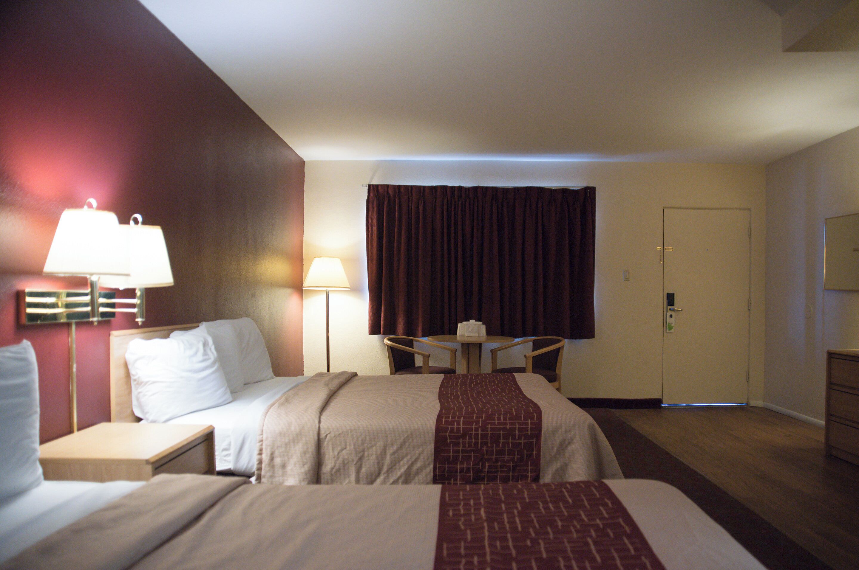 A hotel room with two beds, a desk, a chair, a lamp, a door, and a window with curtains.
