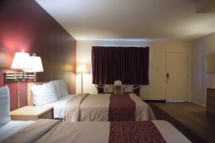 Quarto de hotel com duas camas, uma escrivaninha, uma cadeira, um abajur, uma porta e uma janela com cortinas.