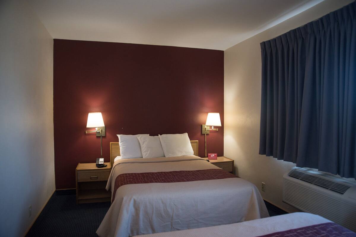 Ein Hotelzimmer mit zwei Betten, einem Nachttisch mit Lampe, einem Heizkörper und einem Fenster mit Vorhängen.