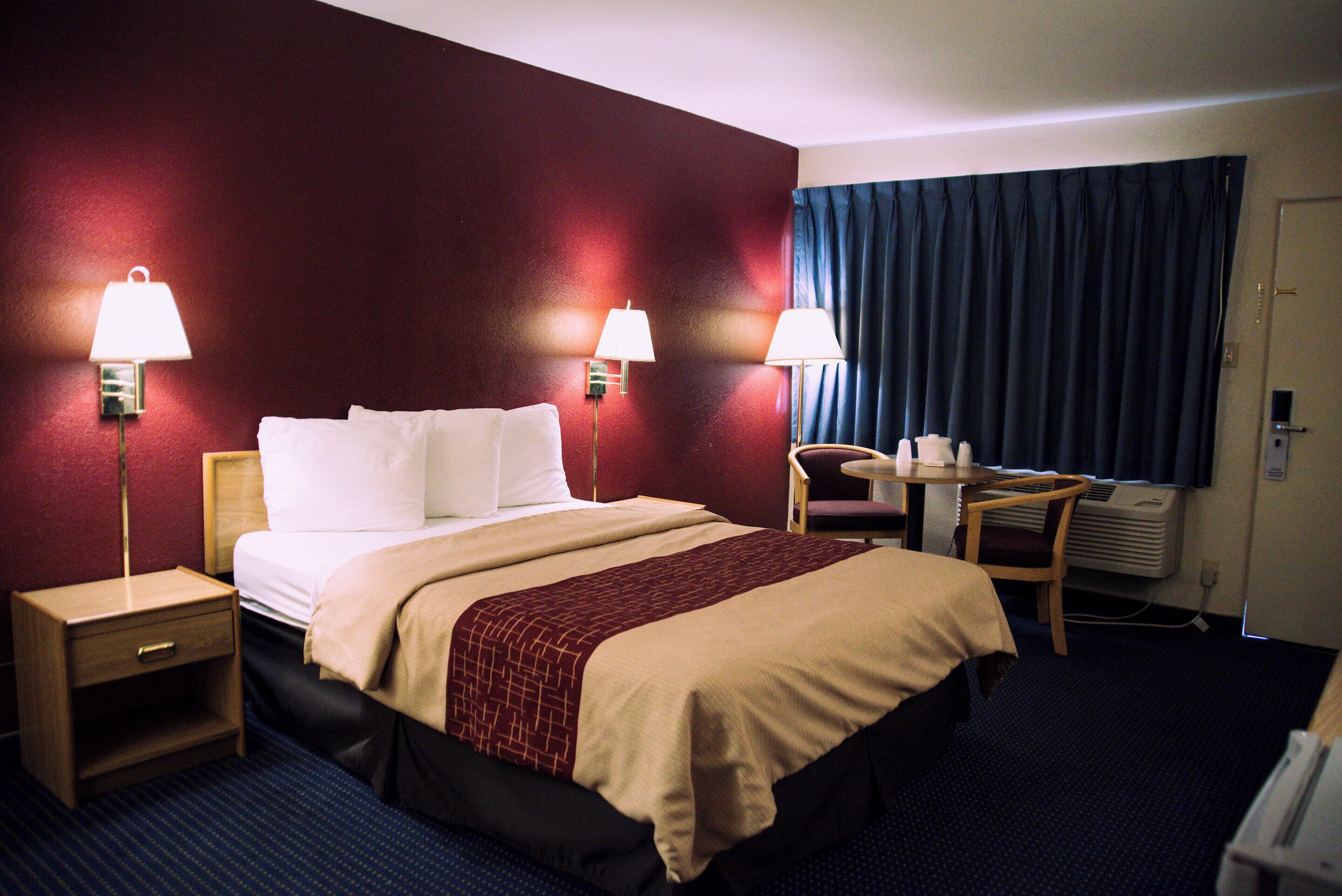 A hotel room with a bed, two bedside lamps, a desk with a chair, and a window with curtains.