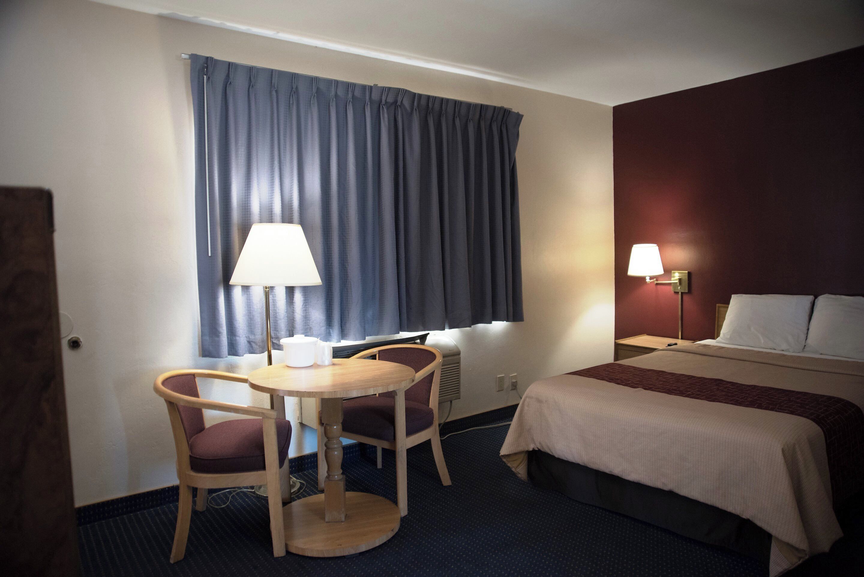 A hotel room with a bed, a desk with two chairs, a window with curtains, and a wall-mounted lamp.