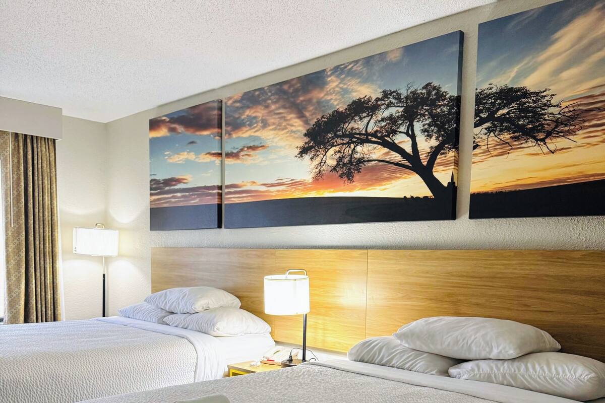Hotellihuone, jossa on kaksi sänkyä, suuri auringonlaskua kuvaava taulu ja yöpöydällä lamppu.