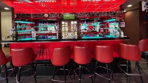 Zona de bar con temática roja, que incluye una barra con estantes llenos de botellas y vasos, además de taburetes rojos.