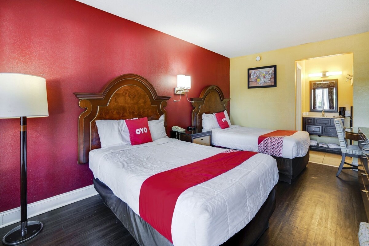 A hotel room with two beds, a desk, a chair, a lamp, and a mirror.
