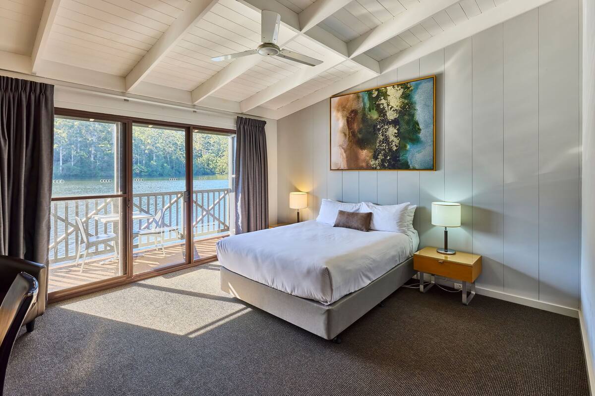 A hotel room with a large bed, a ceiling fan, a painting, and a view of a deck and lake.