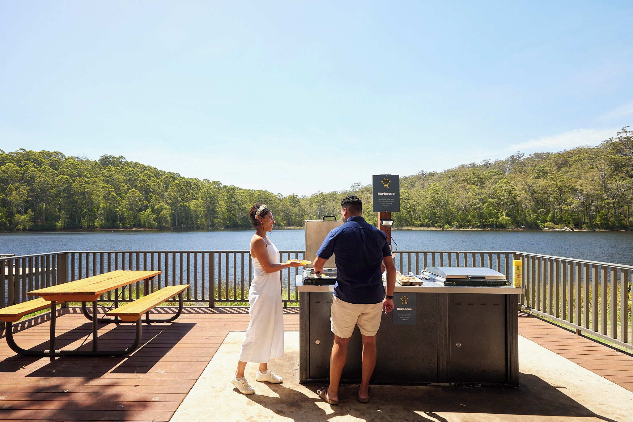 Una terraza de madera con mesas de picnic y una parrilla, con vista a un lago bordeado por un bosque.