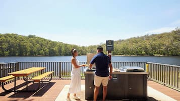 Una terraza de madera con mesas de picnic y una parrilla, con vista a un lago bordeado por un bosque.