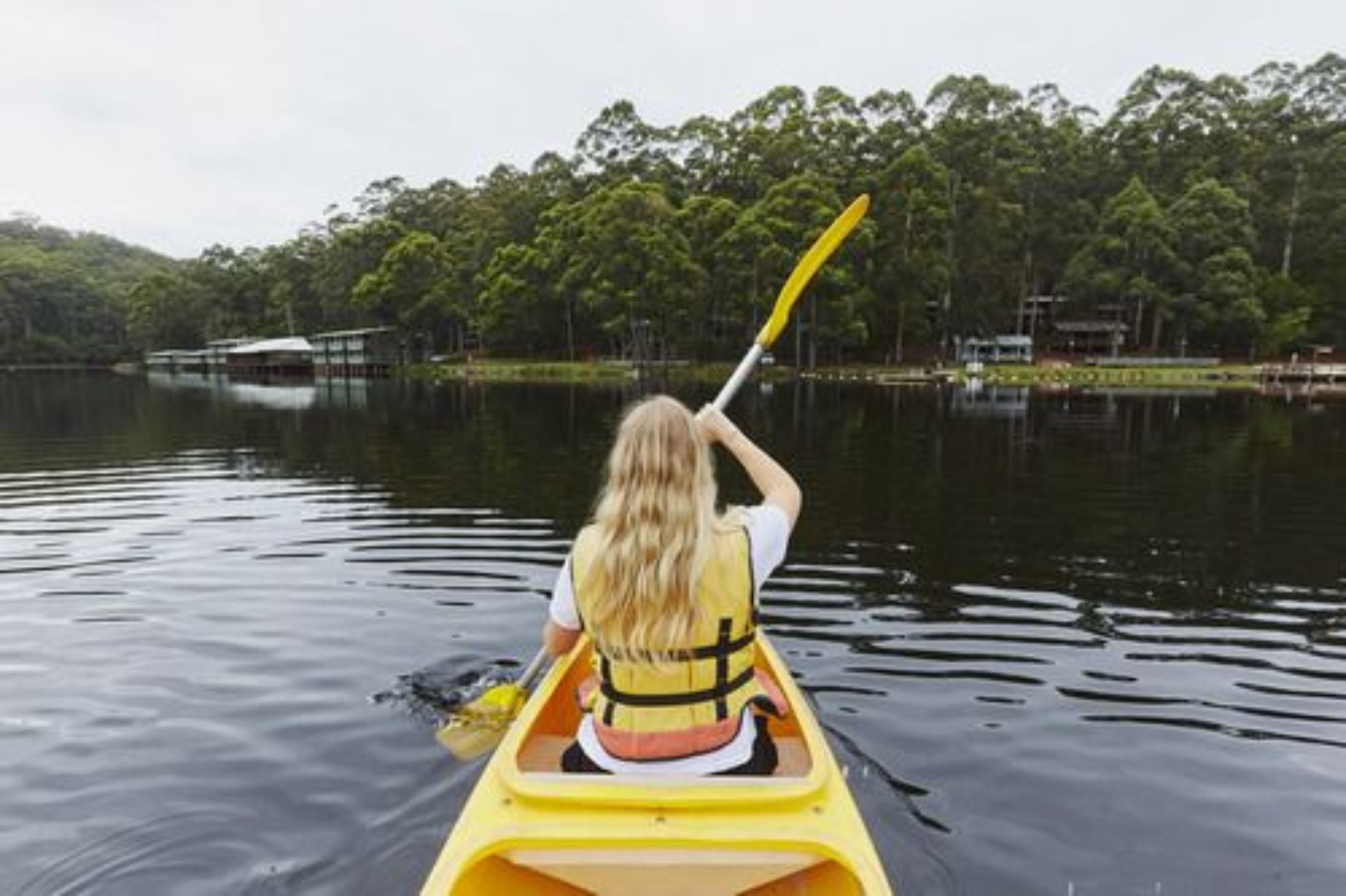 Une personne fait du kayak sur un lac calme, avec une rive boisée en arrière-plan.