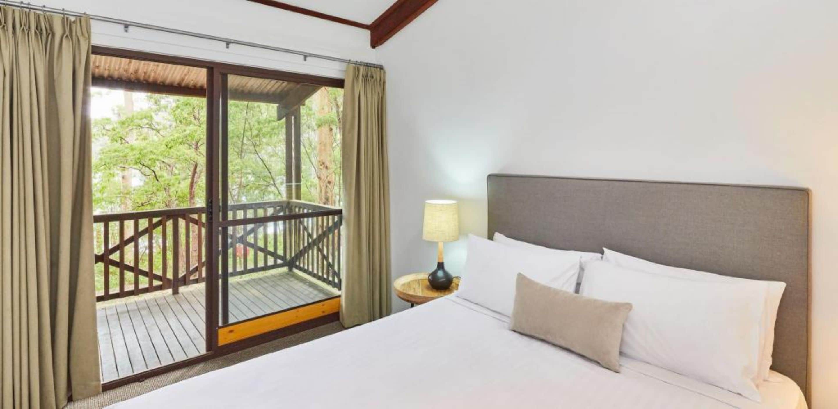 A hotel room with a bed, a nightstand, a lamp, and a balcony with a wooden railing.