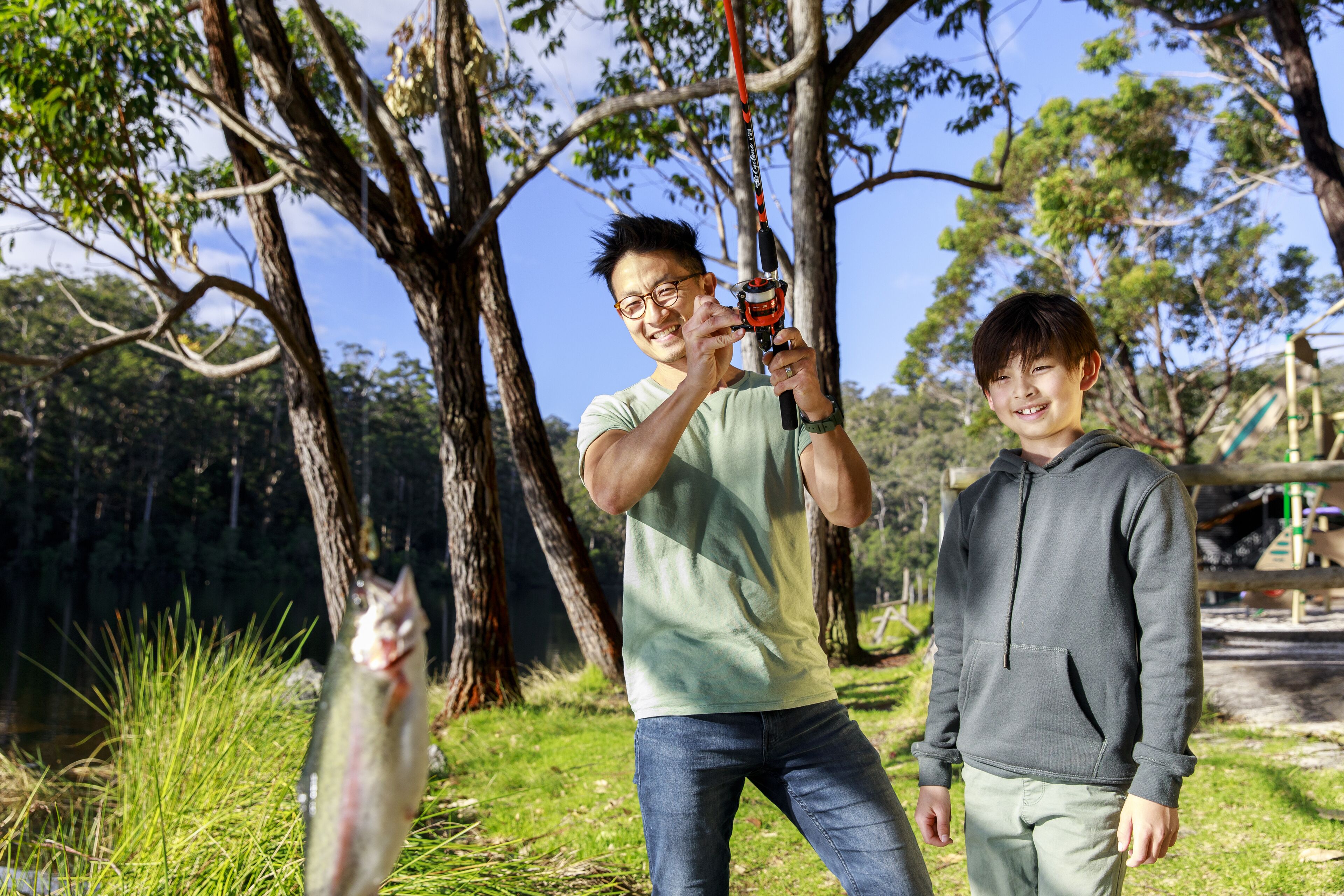 A man holding a fishing rod with a fish, standing next to a young boy in a park with trees and a playground in the background.
