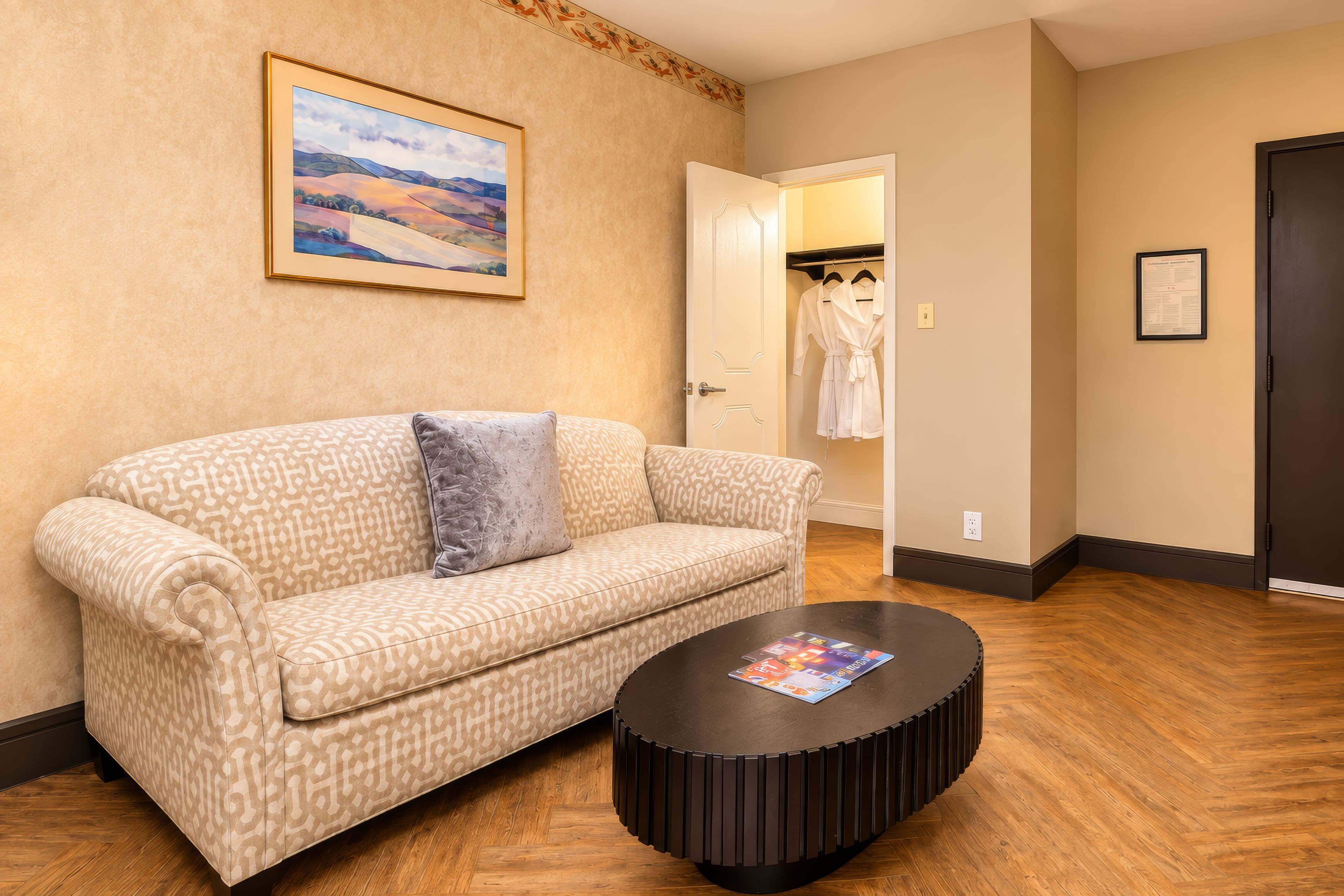 A hotel room with a patterned sofa, a round coffee table, a framed landscape painting, a wardrobe with robes, and a door with a sign.