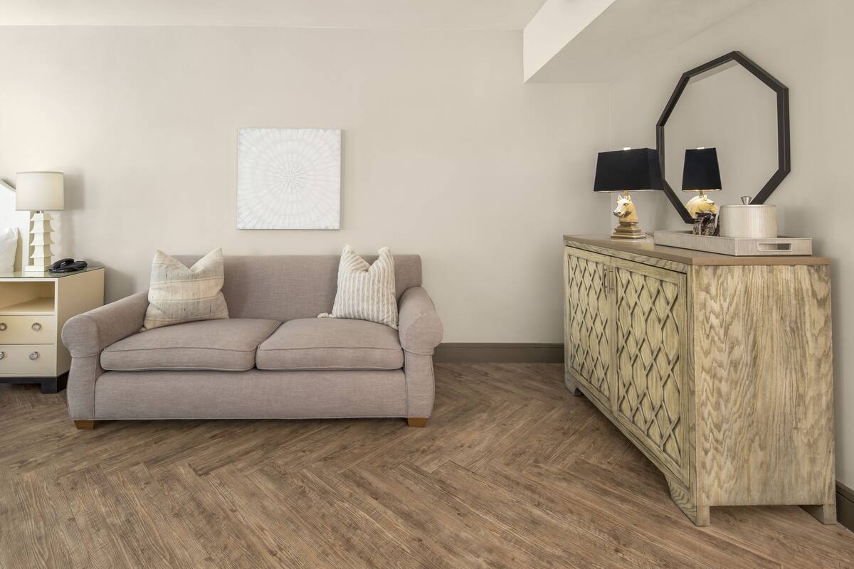 A neutral-toned living room with a sofa, a wooden herringbone-patterned floor, a cabinet with a mirror, and a wall-mounted abstract art piece.