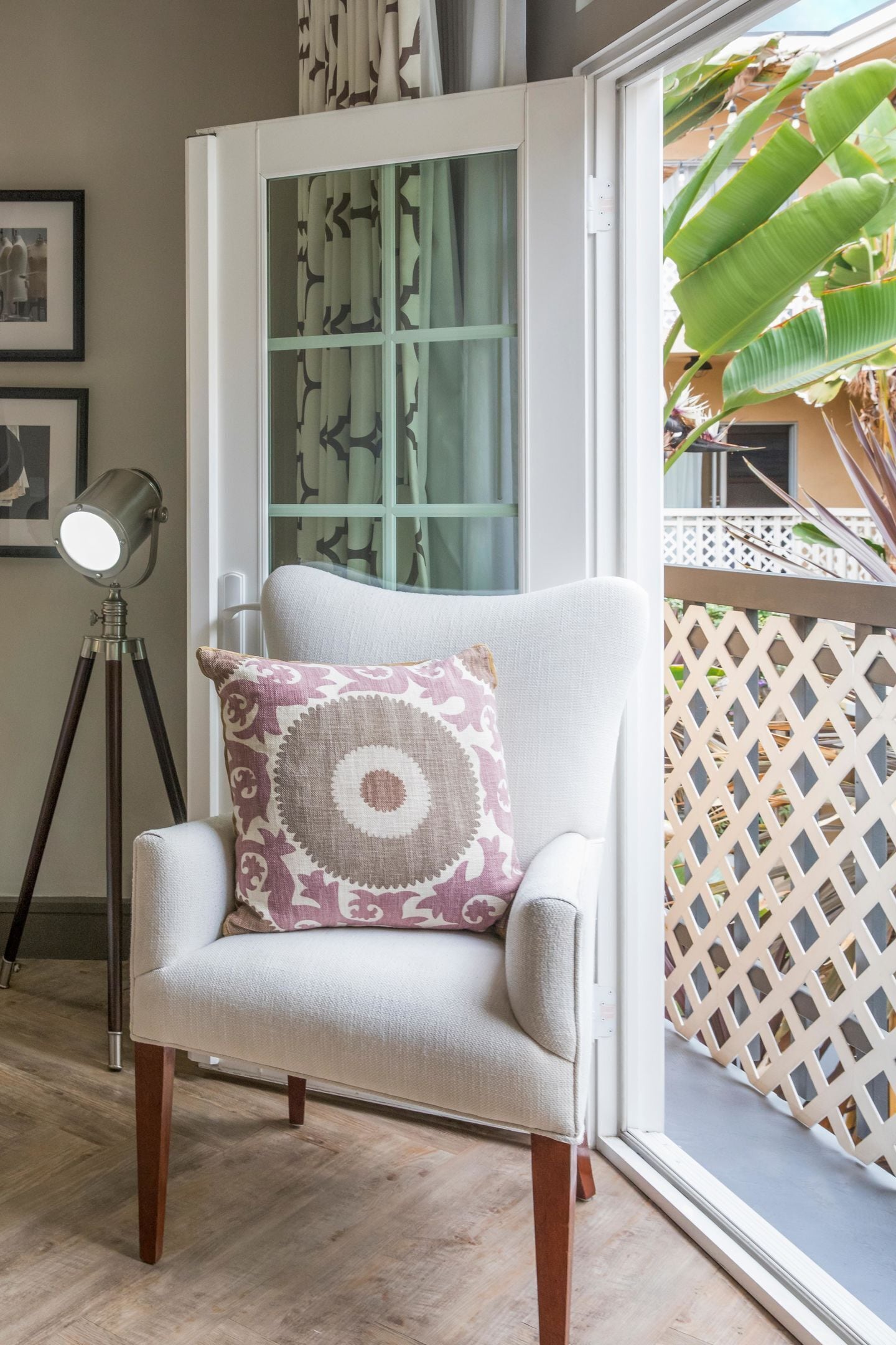Una habitación con un sillón blanco, un cojín estampado, una lámpara de pie y vistas a un balcón con un enrejado y una planta de plátano.