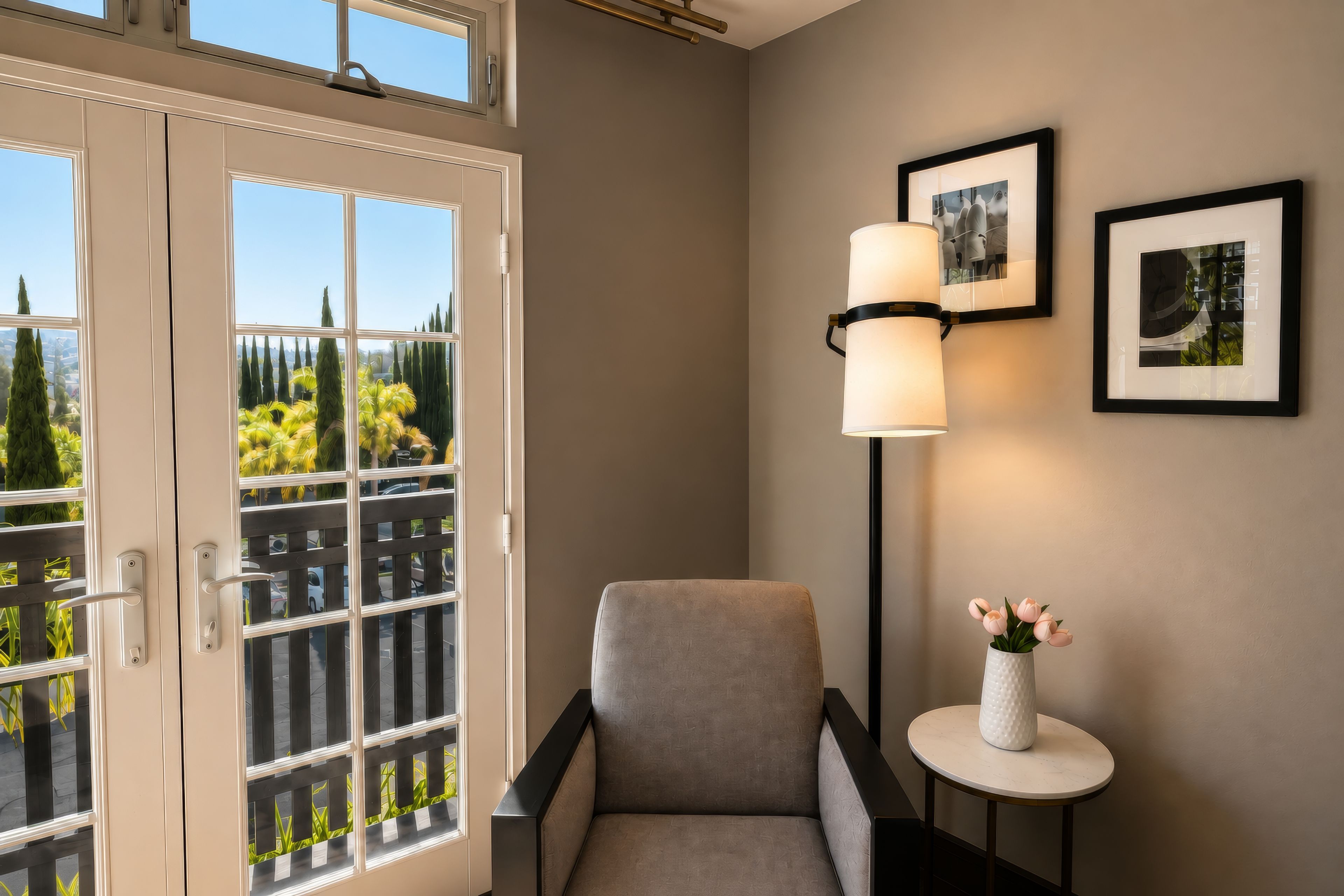 A room with a view of a balcony, a chair, a small table with a vase, and two framed pictures on the wall.