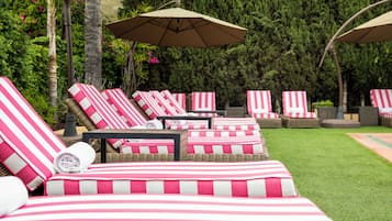 Une rangée de chaises longues rayées, dotées de parasols, est disposée sur une pelouse, avec une haie verte et touffue en arrière-plan.