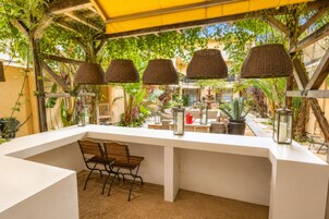 Un'area pranzo all'aperto con tavolo bianco, lampade in vimini e piante in vaso.