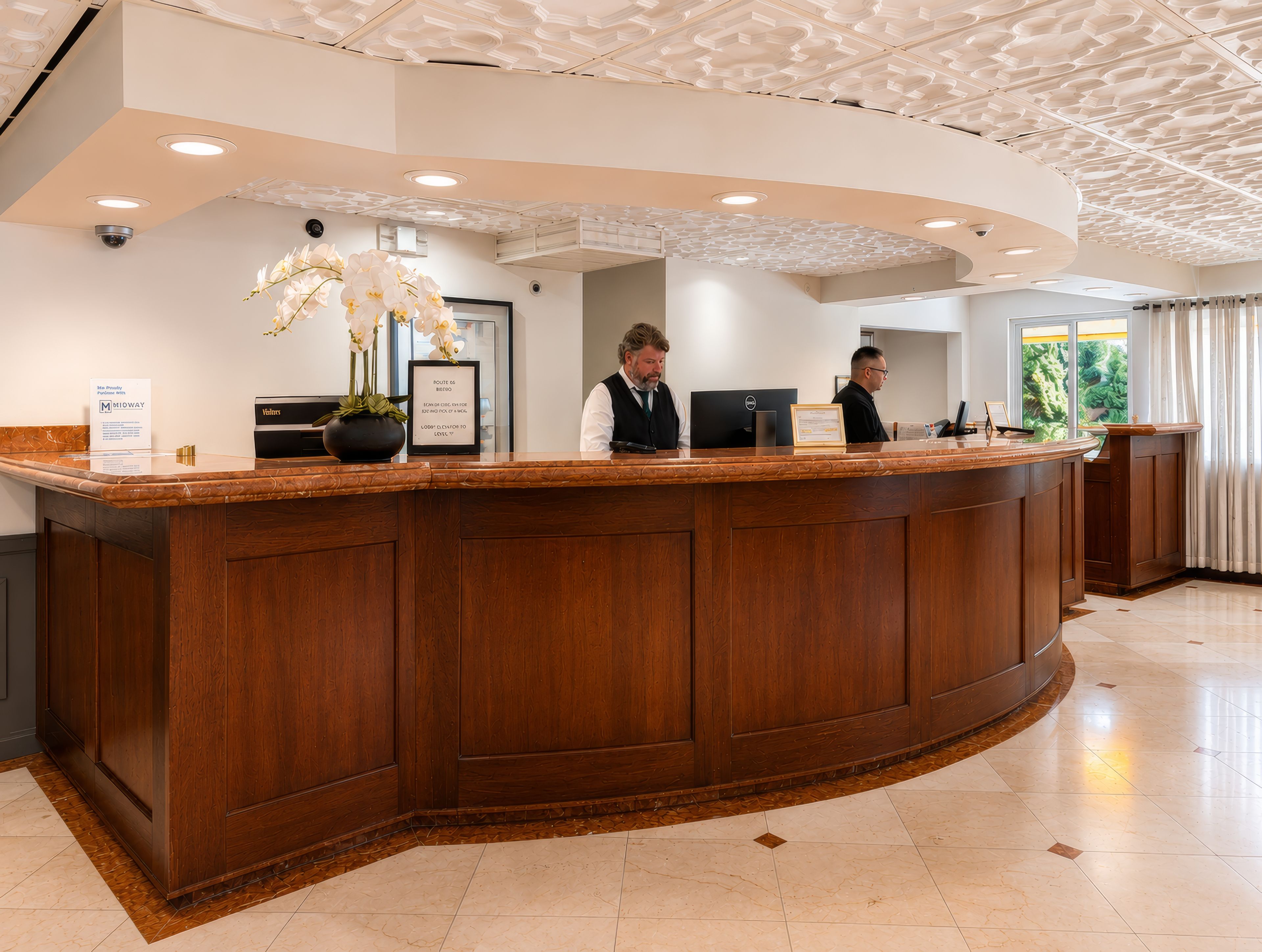 A reception desk with two staff members, a potted orchid, and a framed sign.