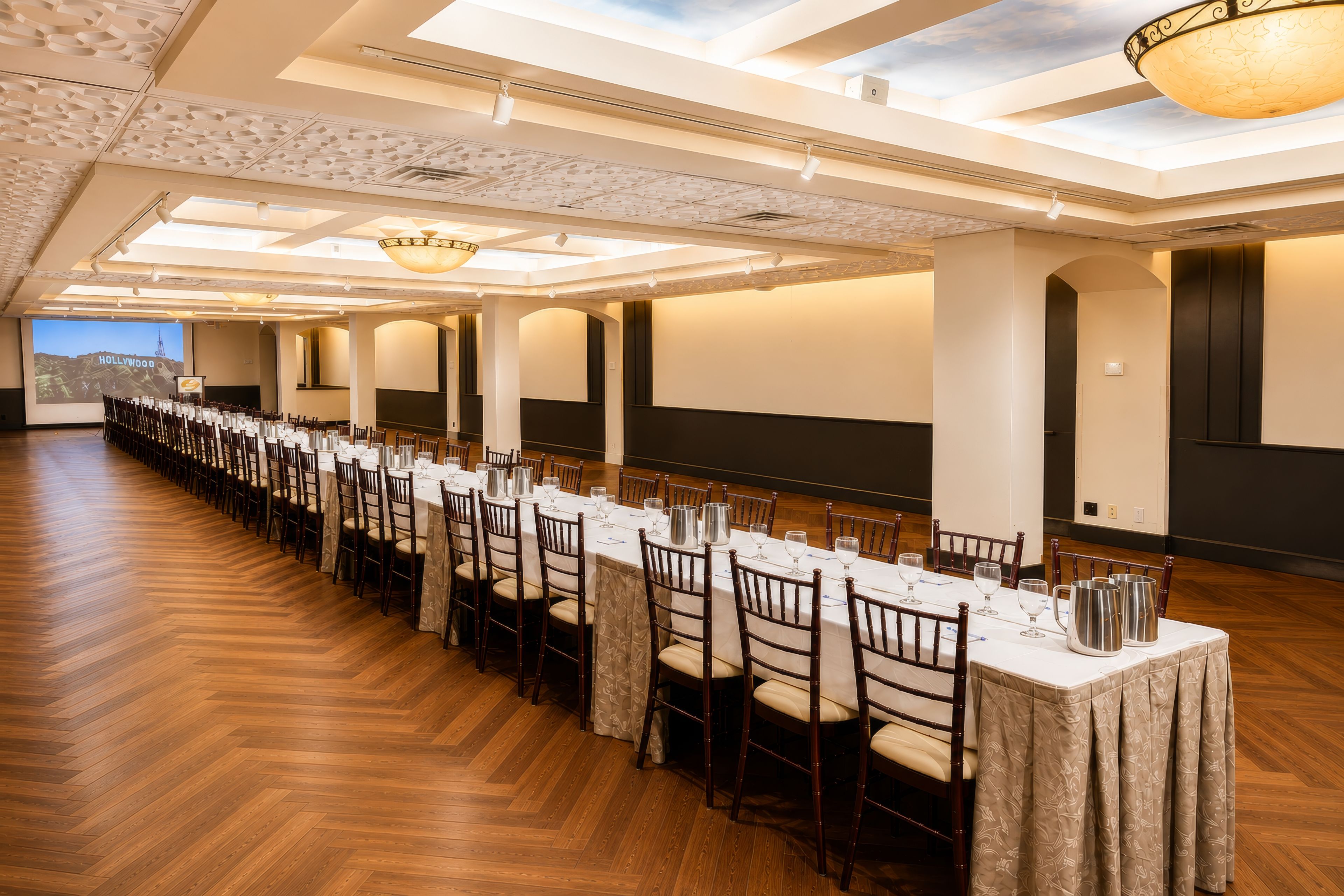 Une longue salle de banquet dotée d’un plancher en bois, de tables et de chaises blanches, et d’un grand écran diffusant une vue panoramique sur la ville.