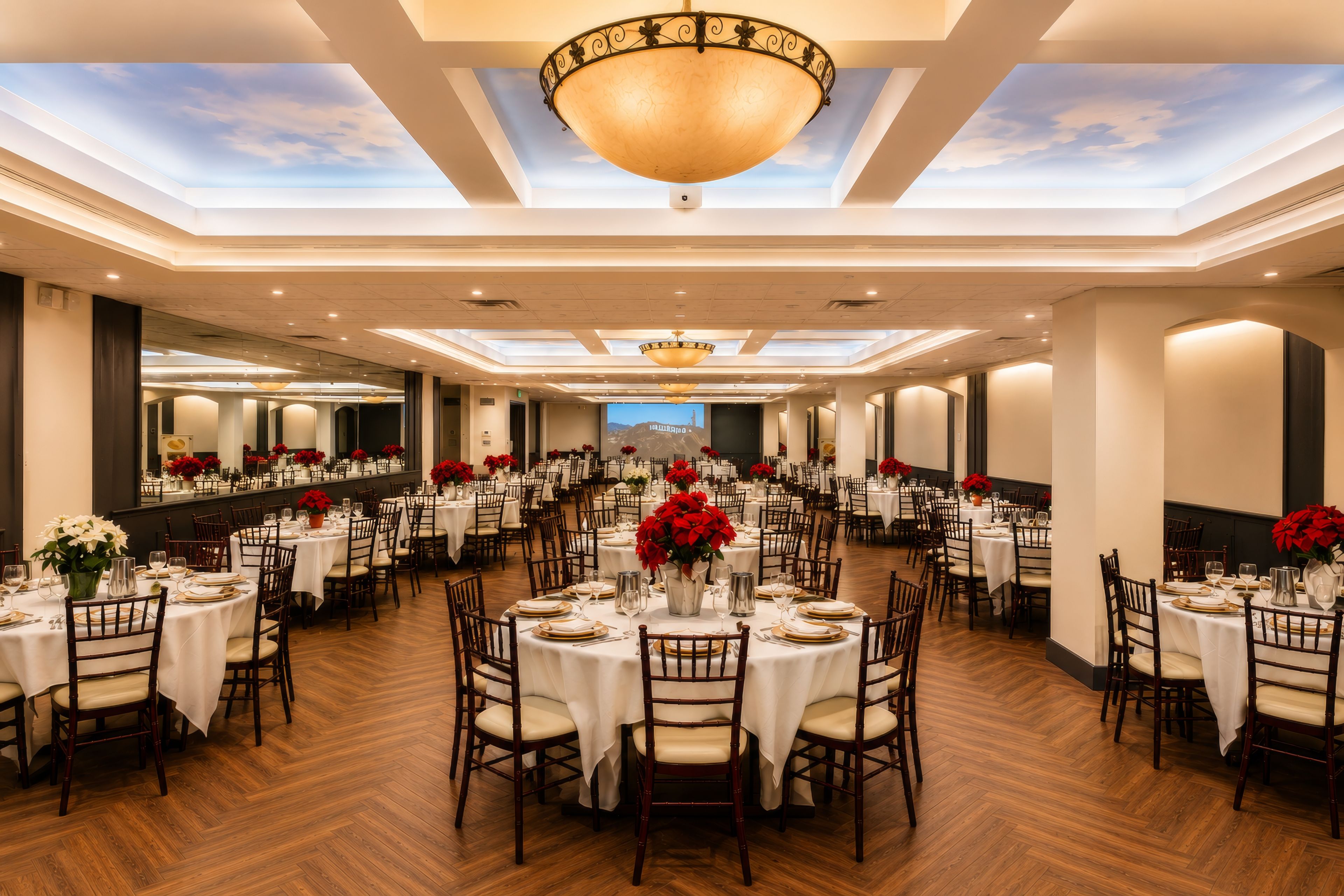 A spacious banquet hall with multiple round tables, each set with white tablecloths and red floral centerpieces.