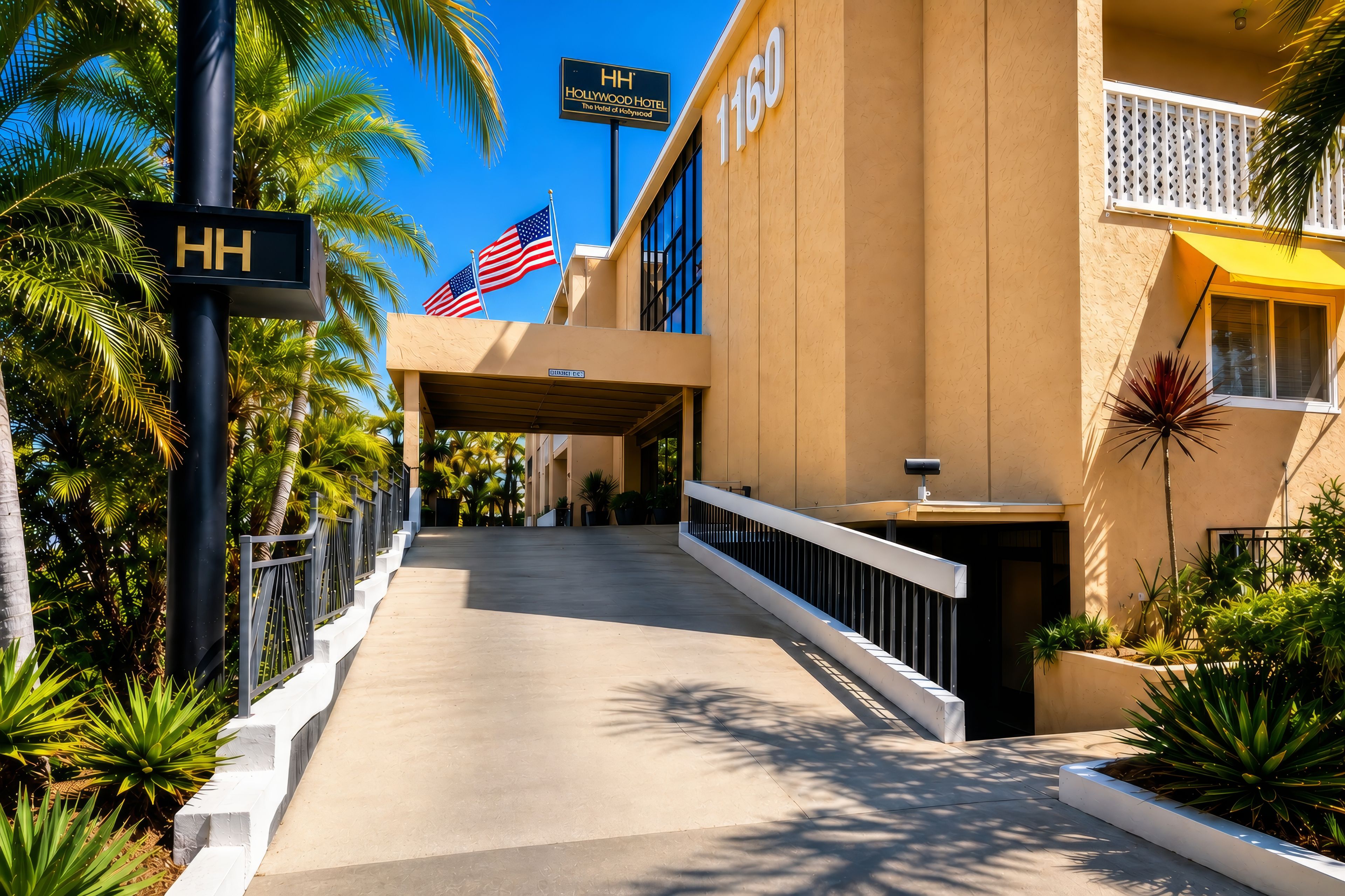 A hotel entrance with a palm tree-lined pathway, an American flag, and a sign for the Hollywood Hotel.
