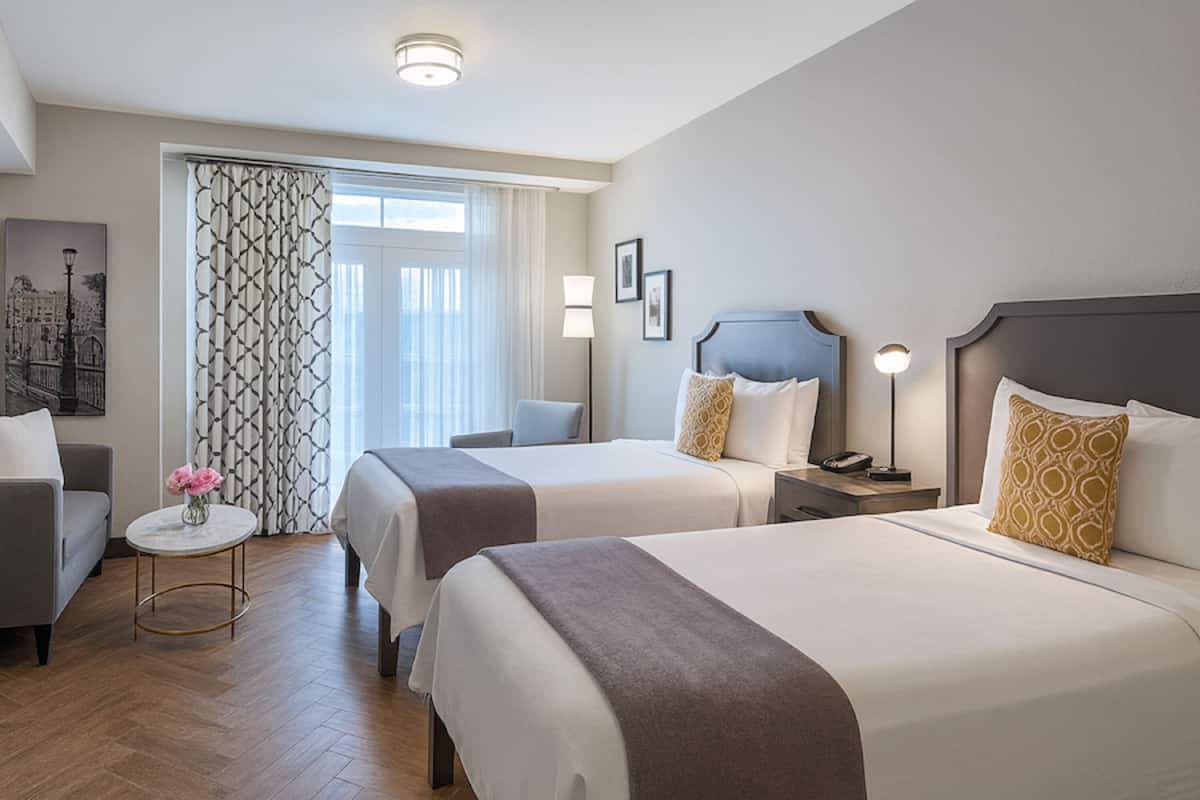 A hotel room with two beds, a sofa, a small table with flowers, and a window with curtains.