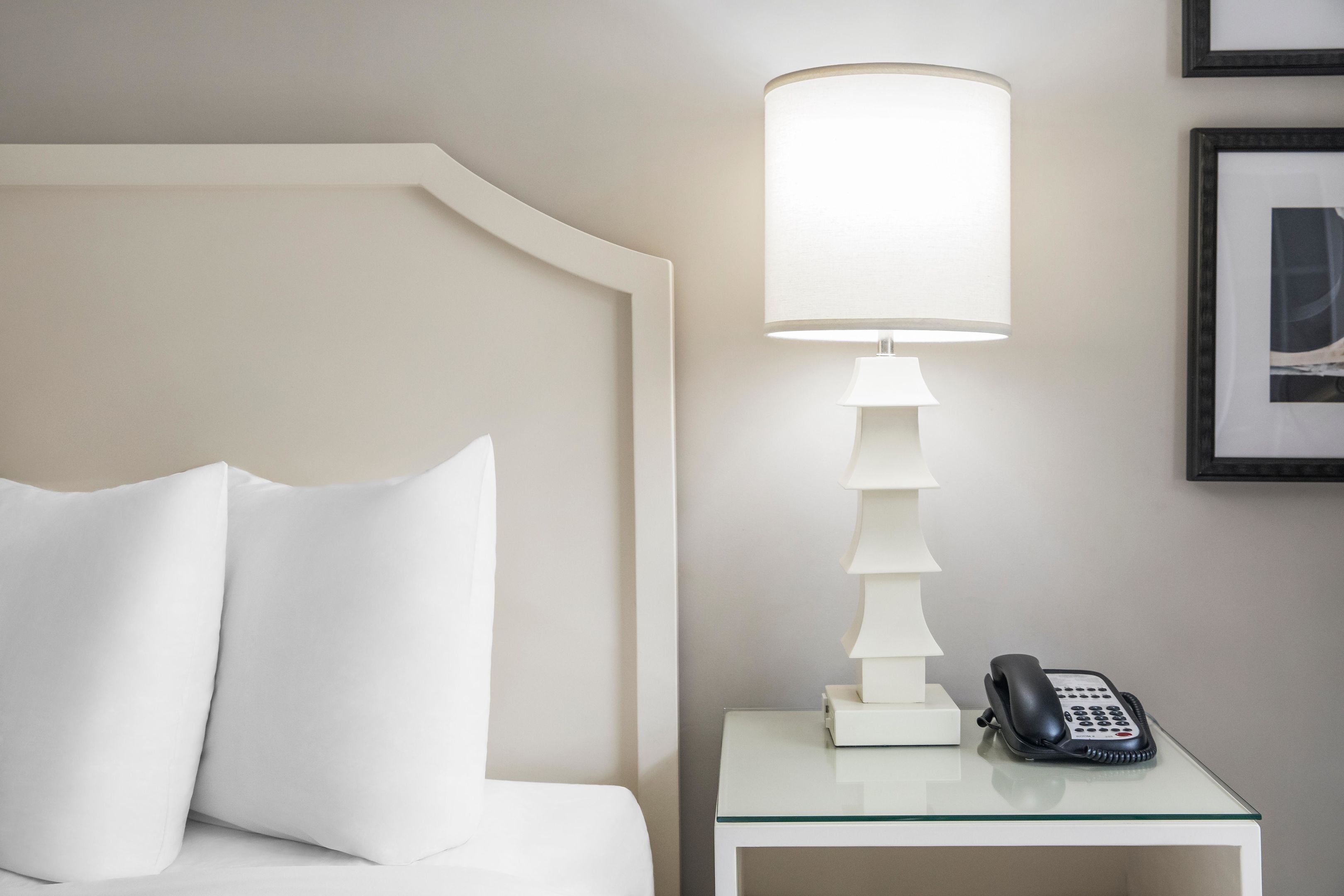 Una cama bien tendida con ropa de cama y almohadas blancas, una mesita de noche con una lámpara y un teléfono, y un cuadro enmarcado en la pared.