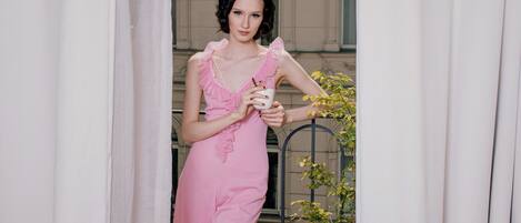 Une personne vêtue d’une robe rose, debout dans un doorway, avec un rideau blanc.