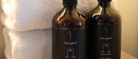 Two bottles of Saint Charles Hotel Motto Wild Roots Hand & Body Lotion on a wooden surface with a white towel in the background.