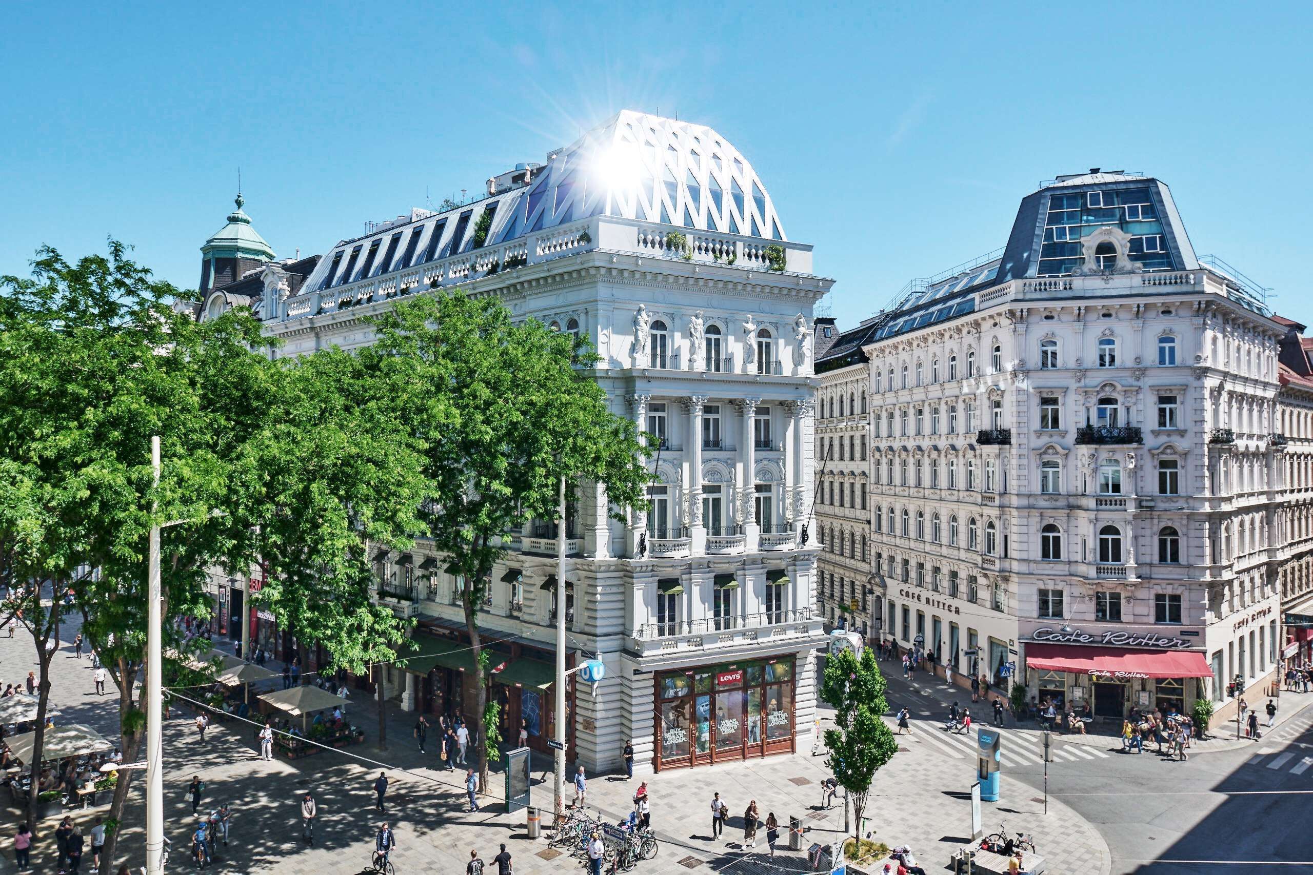 Um edifício histórico com arquitetura clássica, um café no andar térreo e uma praça pública movimentada com árvores e pedestres.