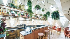 A modern bar area with a wooden bar counter, bar stools, and a variety of plants hanging from the ceiling. - Hotel MOTTO (Vienna)