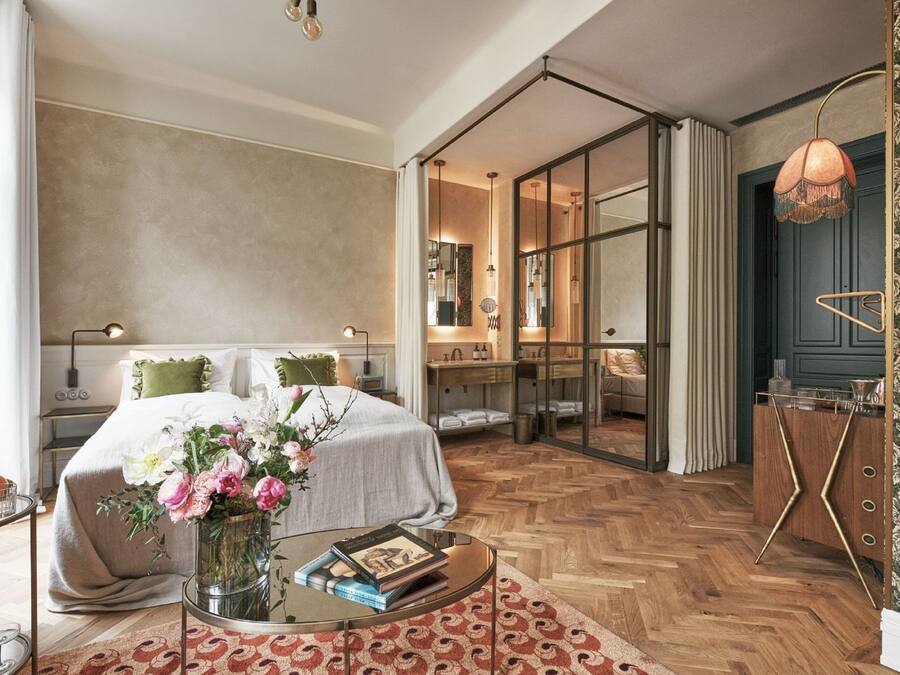 Una habitación de hotel moderna con una cama grande, un baño con paredes de vidrio, piso de madera con diseño de espiga y una pequeña mesa redonda con un jarrón de flores.