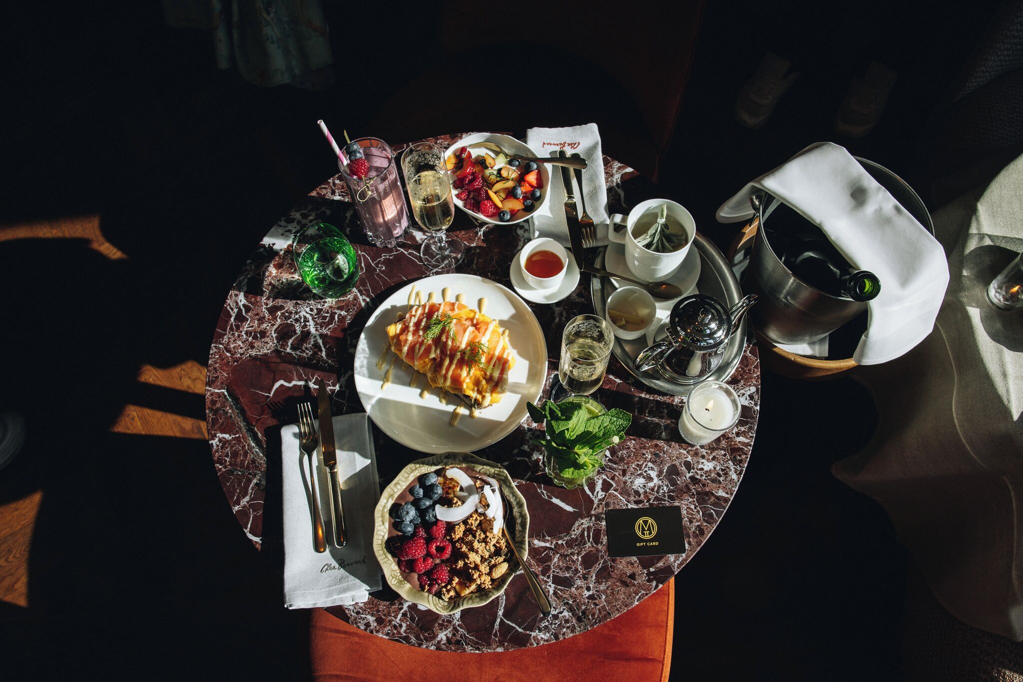 A dining table with a variety of dishes, including a plate of fruit, a bowl of granola, and a dessert. There are also several glasses, a cup, and a bottle on the table.