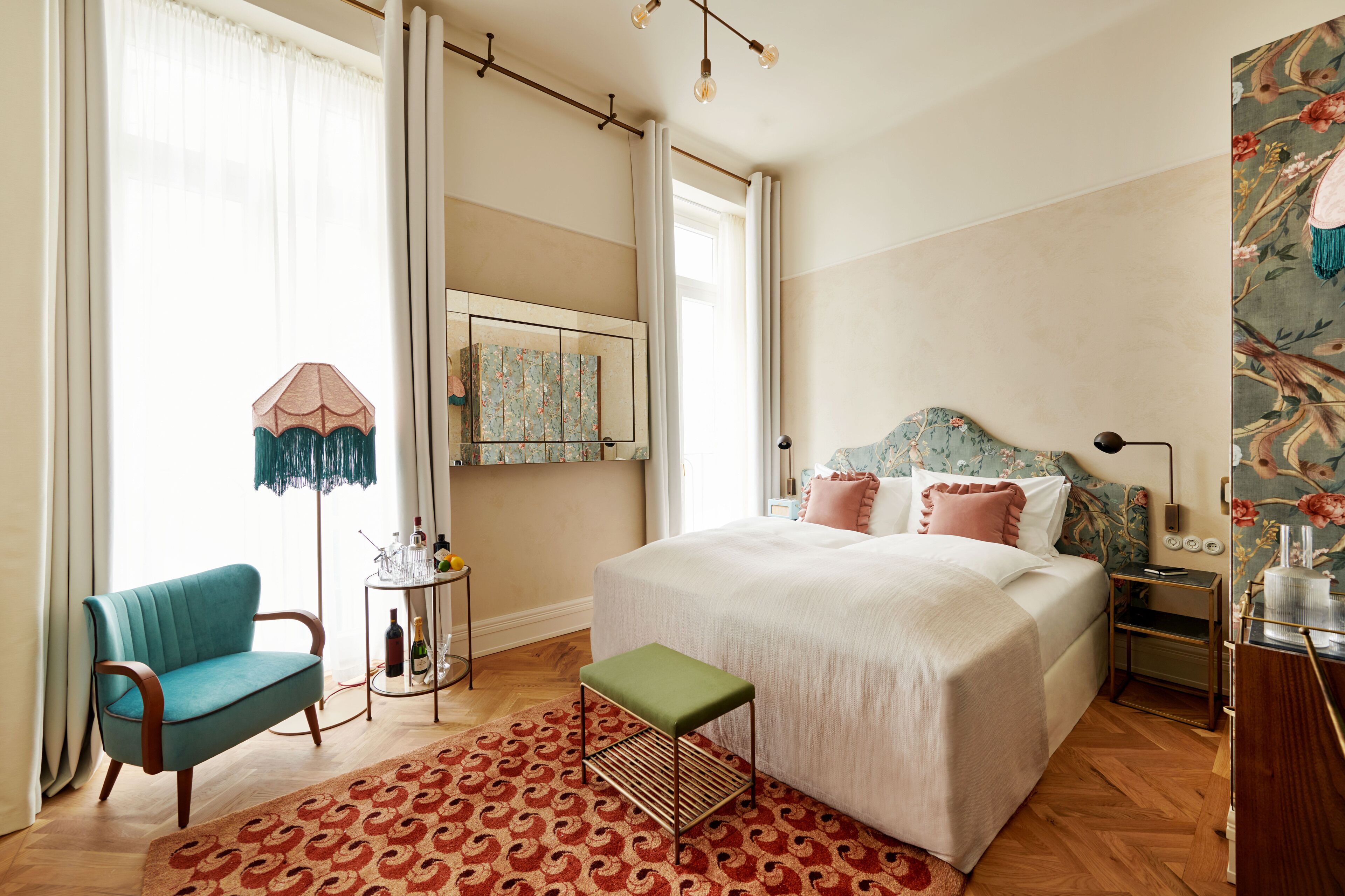 Une chambre avec un grand lit, un fauteuil vert, une petite table avec des bouteilles et un tapis à motifs.